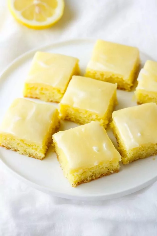 These lemon brownies are presented as neatly cut squares with a vibrant yellow hue, indicating a moist and tender texture. The base layer appears soft and cake-like with a slightly dense crumb, while the top is coated with a smooth, glossy lemon glaze that has a slightly translucent quality.